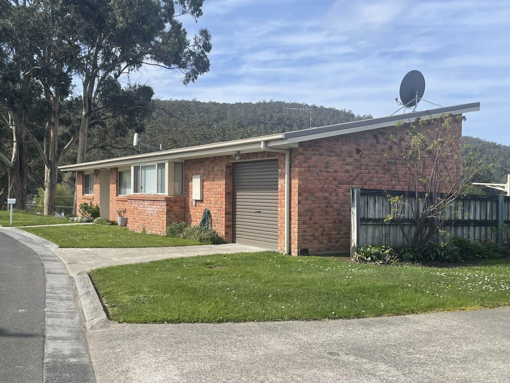 Single-story brick house with a sloped roof, garage door, and satellite dish. It is surrounded by a small lawn, driveway, and wooden fence, with trees and hills in the background under a partly cloudy sky.