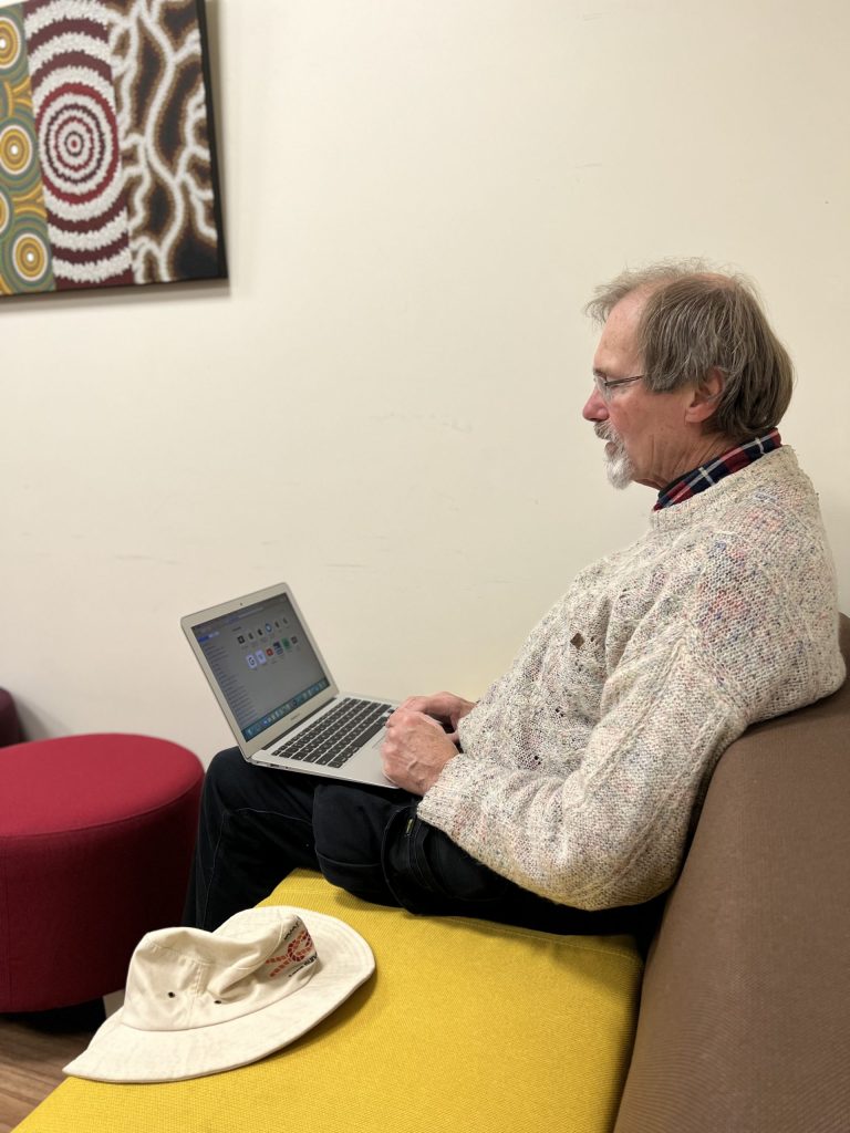 An elderly man with glasses and a beard is sitting on a bench, typing on a laptop. Hes wearing a gray sweater and dark pants. A hat is placed next to him. A colorful abstract painting hangs on the wall behind him.