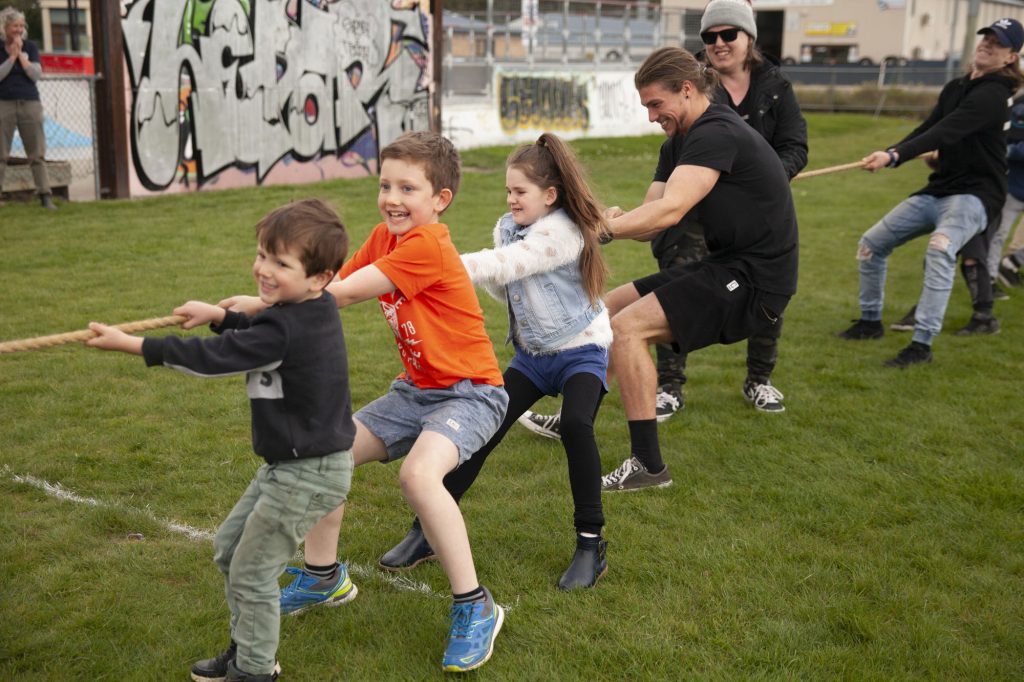 A group of people, including children and adults, are playing tug-of-war on a grassy field. The background features graffiti-covered walls and some buildings. Participants are smiling and appear to be enjoying the game.