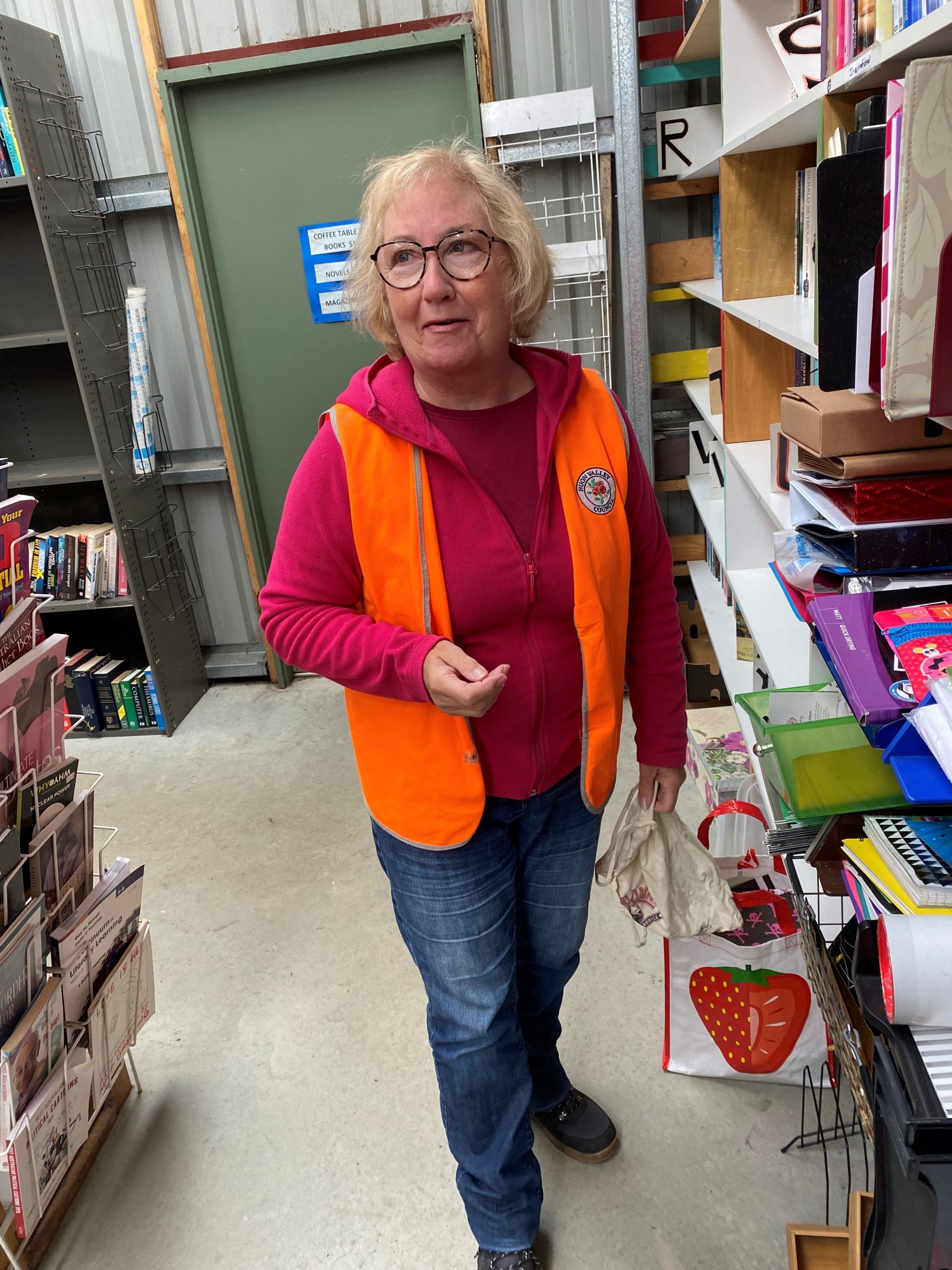 Elderly woman with glasses, wearing an orange vest and red sweater, walks through a store aisle beside shelves with books and stationery items. Shes carrying a shopping bag with a strawberry design.