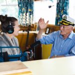 A woman wearing headphones holds a microphone toward a man in a captains hat, who gestures with his hand. They are sitting inside a boat or nautical-themed room, with decorative model ships and a bowl of fruit visible in the background.