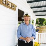 An older man wearing a captains hat and blue shirt stands smiling on a white porch. He is near a door with a wooden sign that reads Stella. In the background, there is greenery and water.