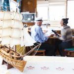 A detailed wooden ship model with white sails is on a table in the foreground. In the background, a man and a woman are sitting and talking in a cozy, sunlit room, with a microwave visible behind them.