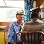 A man in a captains hat and blue shirt is smiling while sitting at a kitchen table, being interviewed by a woman wearing headphones. The setting appears cozy and bright, with warm wooden interiors and a window with curtains.