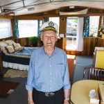 Elderly man wearing a captains hat stands smiling in a warmly decorated room with nautical accents. A bed with a blue-patterned cover is in the background, along with a cozy stove and a table with chairs. Large windows let in ample light.