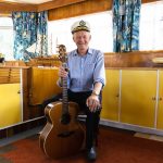 An older man wearing a captains hat sits in a room with yellow cabinets and blue curtains, holding an acoustic guitar. A model ship and a vintage car decoration are on shelves behind him.