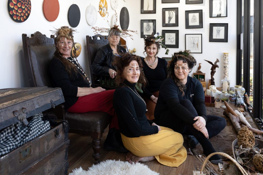 Five women wearing nature-inspired headpieces and face paint are sitting in a room adorned with framed artwork and natural elements. They are seated on chairs and the floor, surrounded by items like logs and a vintage chest, near a well-lit window.