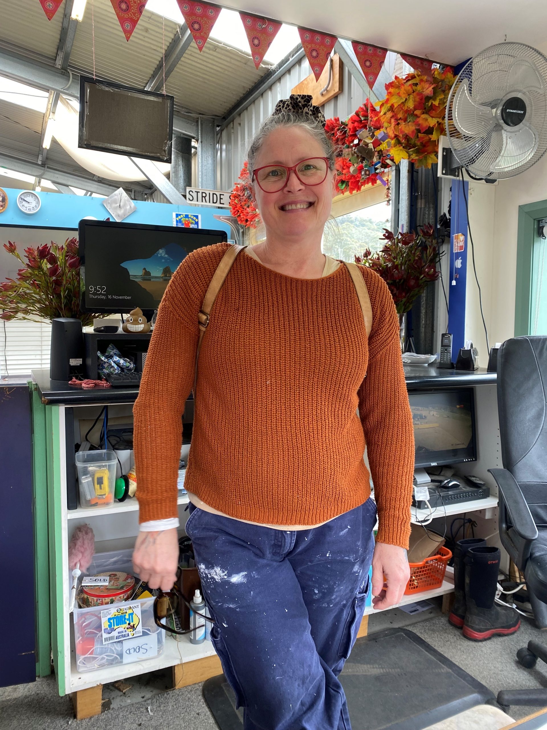 A person wearing glasses and a brown sweater stands in a cluttered room with a computer, red boots, and autumn decorations. There are paint stains on their pants. They are smiling at the camera.