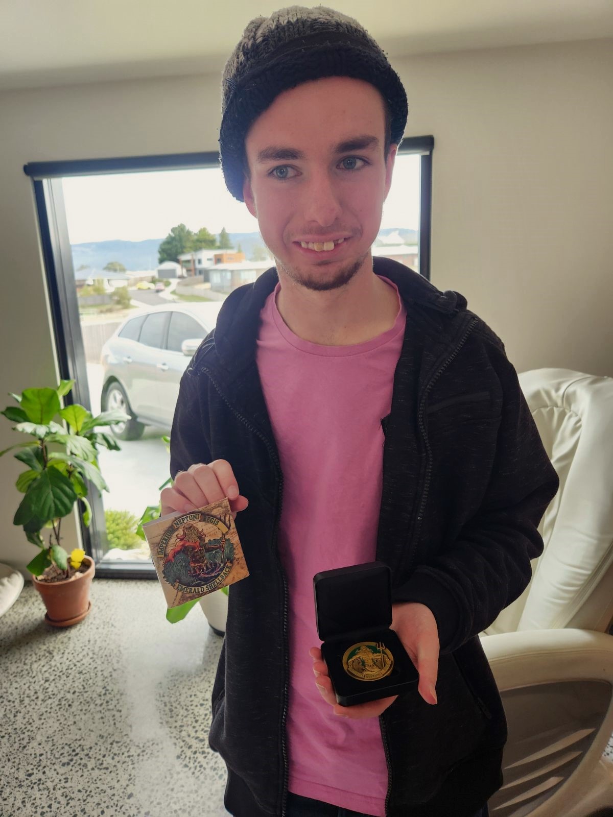 A man in a winter hat and purple shirt holds a commemorative coin in a black box and a colorful card. Hes indoors, standing by a window with a view of a parked car and greenery outside. A potted plant sits in the corner of the room.