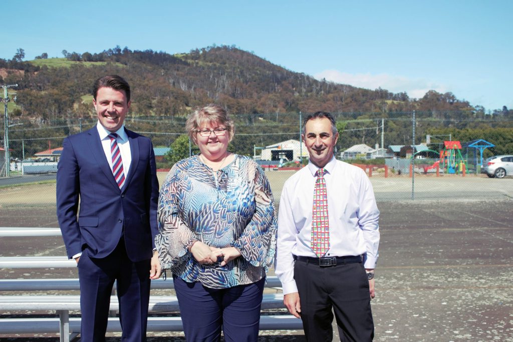 Three people are standing outside on a paved area with a playground and hills in the background. The person on the left is wearing a suit, the middle person is in a patterned blouse, and the right person is in a white shirt with a patterned tie.