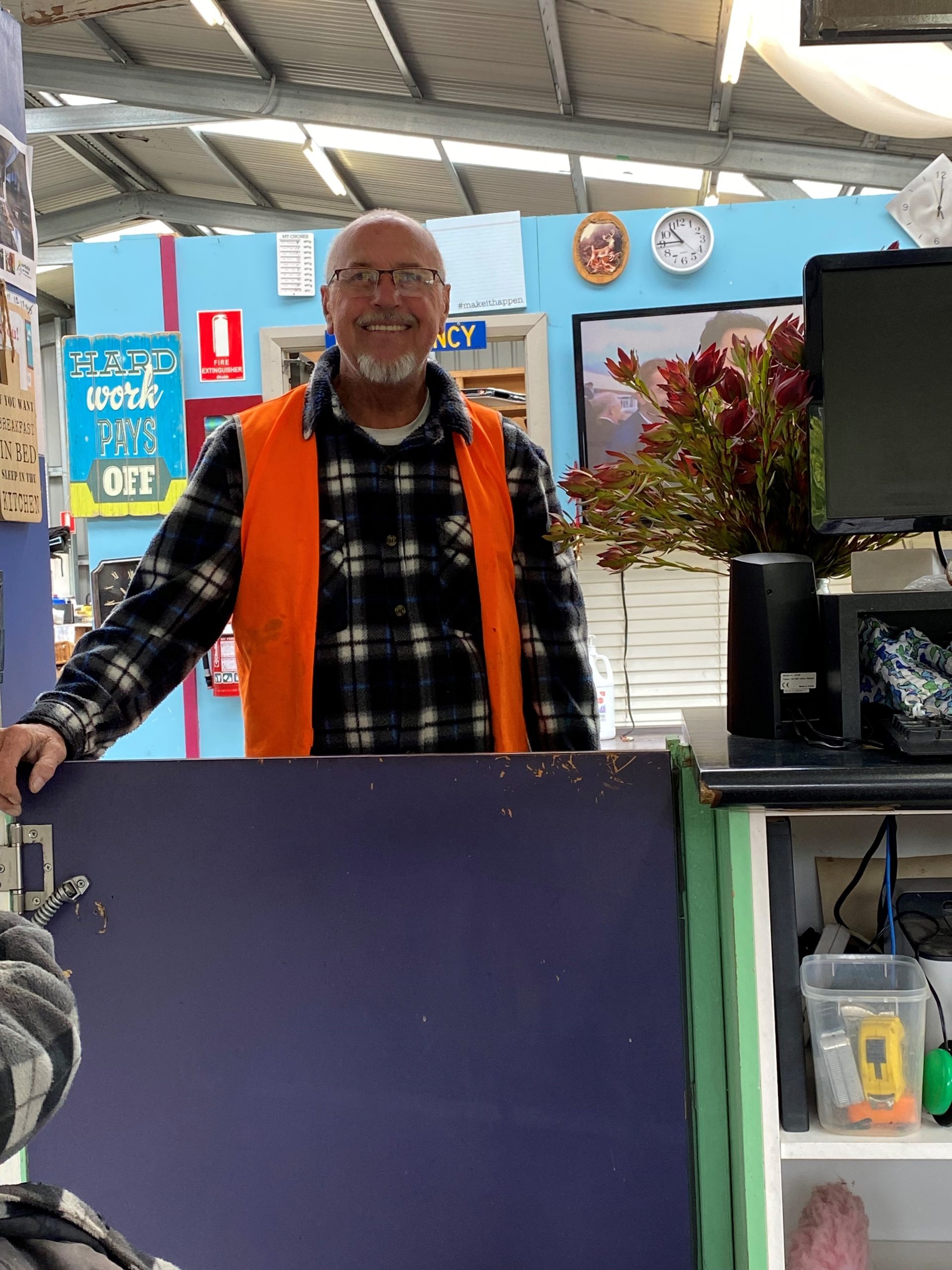 A man wearing glasses, an orange safety vest, and a plaid shirt is smiling behind a half-door in a brightly colored room. Theres a clock on the wall, signs, and a bouquet of red flowers on a counter nearby.