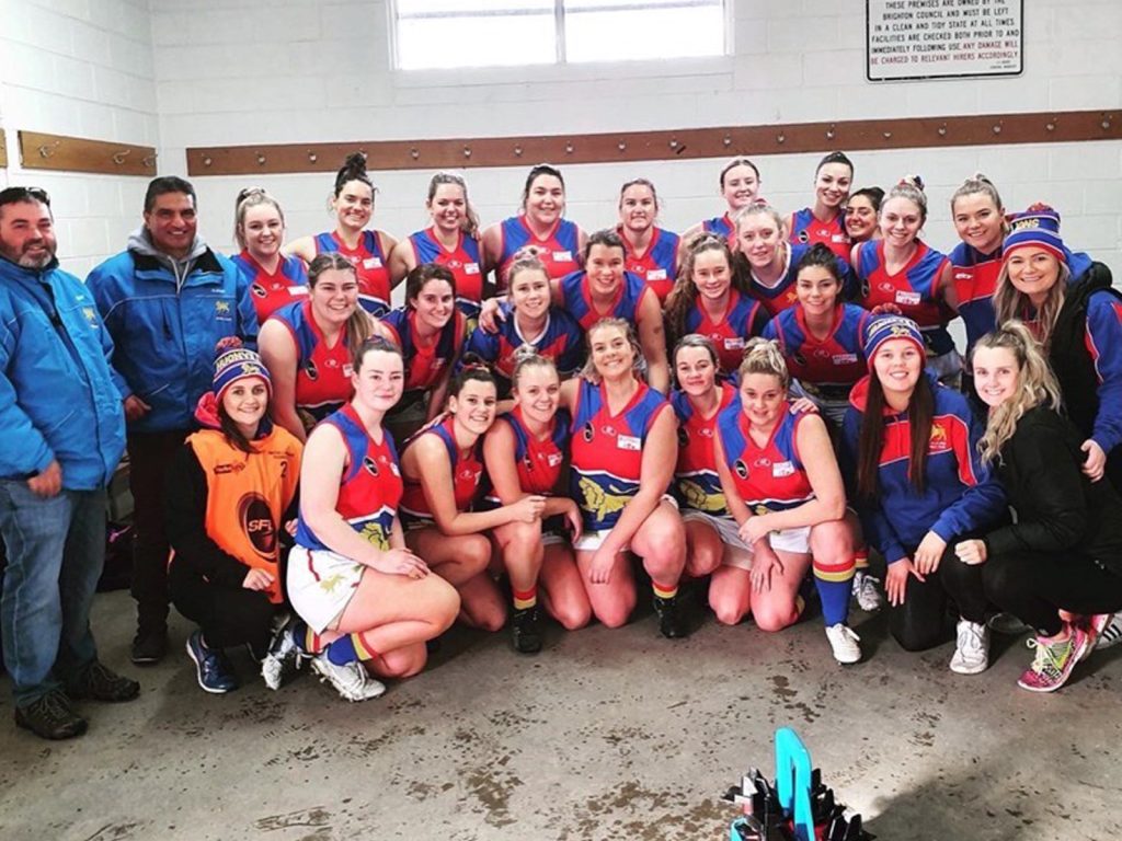 A group of women in matching athletic uniforms and a few men in casual jackets pose together in a sports locker room. The team is smiling and appears to be in high spirits. A window is in the background.