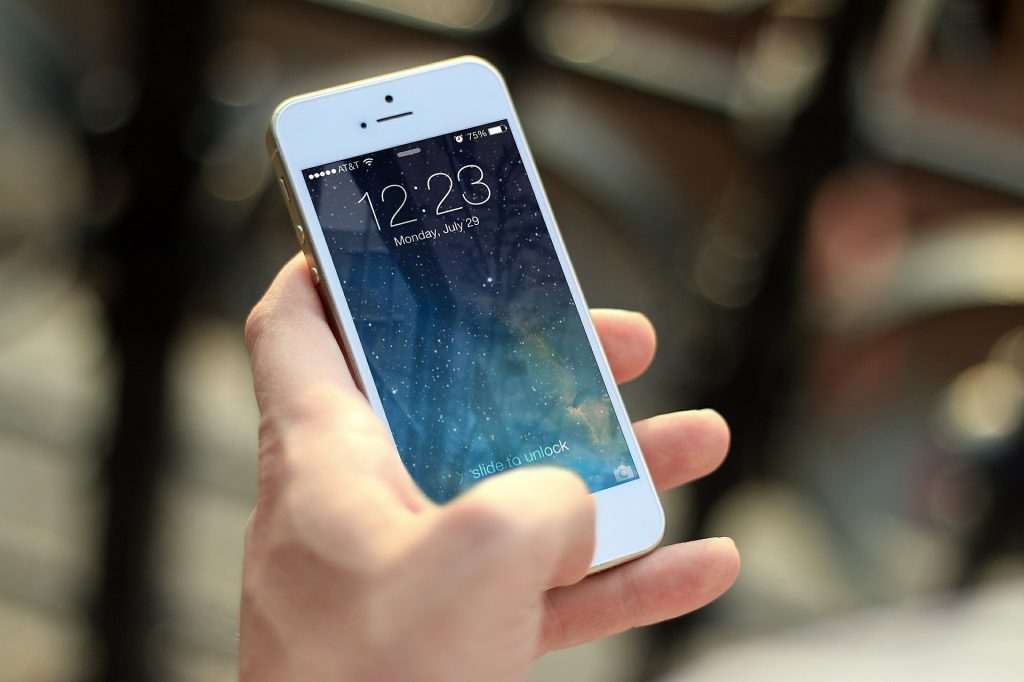 A person holding a white smartphone with the lock screen showing the time as 12:23 on Monday, July 29. The screen displays a slide to unlock prompt. The background is blurred, focusing on the phone.
