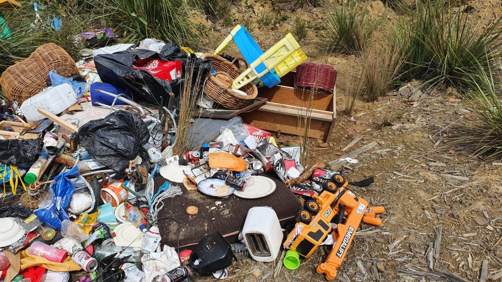 A pile of discarded items, including plastic containers, toys, wicker baskets, and electronics, is scattered on the ground near tall grass. Visible in the heap is an orange toy truck, a toaster, and a yellow object among the debris.