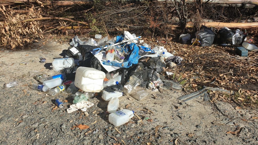 A pile of scattered garbage, including plastic containers, bags, and bottles, sits on a dirt and gravel area surrounded by dry leaves and branches. Plastic bags and other debris are visible in the background.