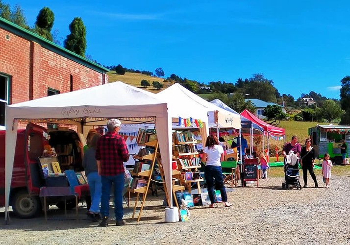 Outdoor market scene with various stalls under white canopies displaying books and other items. People browse and walk around on a sunny day. A backdrop of rolling hills and trees under a clear blue sky completes the setting.