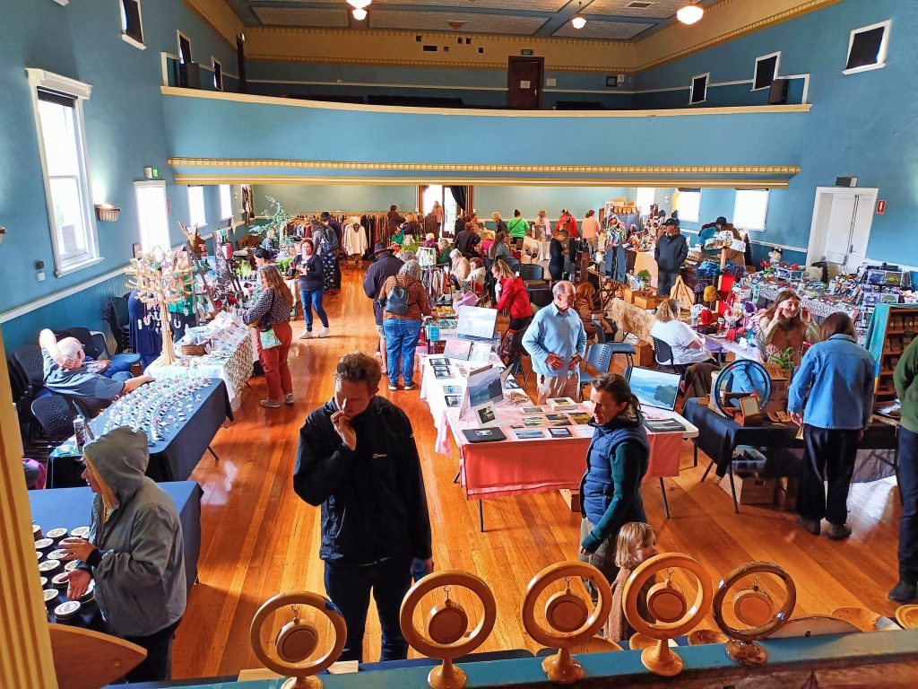 A bustling indoor market is set up in a blue-walled hall with wooden floors. Vendors display crafts, art, books, and clothes on tables, while shoppers browse and mingle throughout the spacious room.