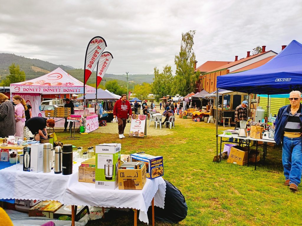 A lively outdoor market with colorful tents and stalls selling various items, including kitchen appliances and food. People are browsing, and there are mountains and trees in the background under a cloudy sky.