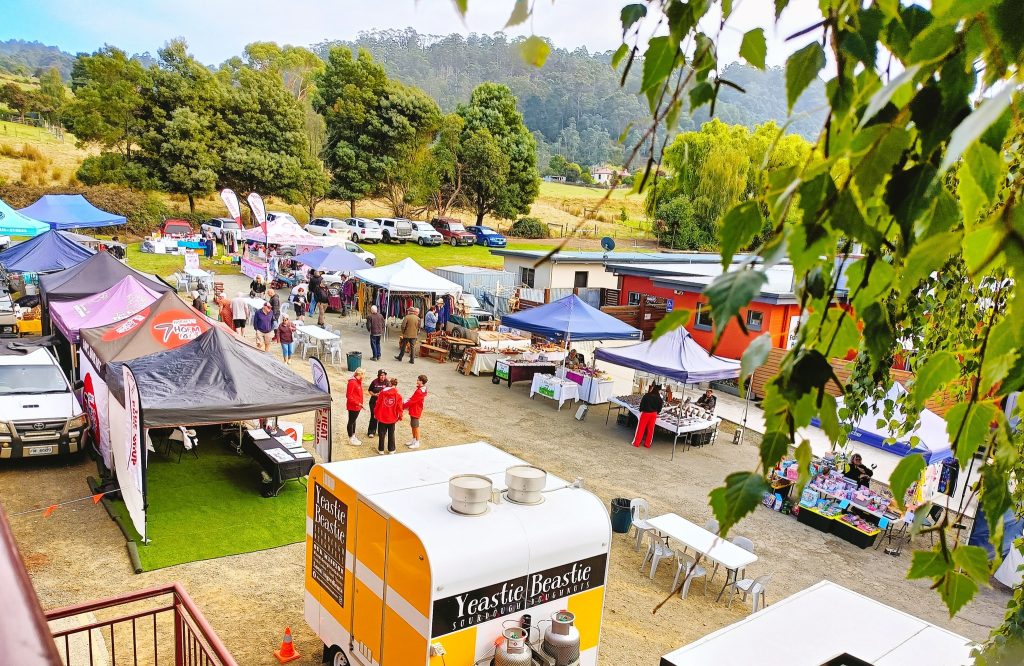 A vibrant outdoor market scene with colorful tents and stalls, people browsing, and a yellow food truck labeled Yeastie Beastie. Surrounded by trees and parked cars, with hills visible in the background.
