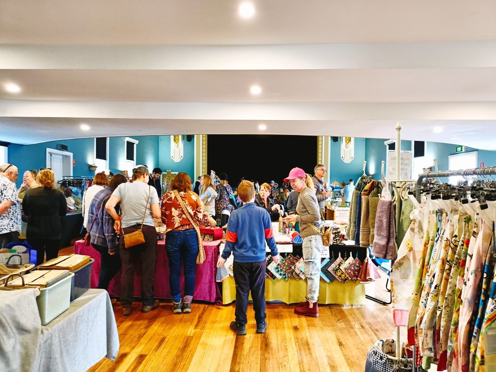 A group of people browse handmade crafts and clothing at an indoor market with wooden floors, colorful tables, and racks of garments. The atmosphere is lively and bright.