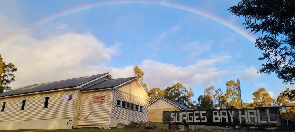 A rainbow arcs across a partly cloudy sky above a cream-colored building labeled Surbges Bay Hall, surrounded by trees, with a large wooden sign in front of the hall.
