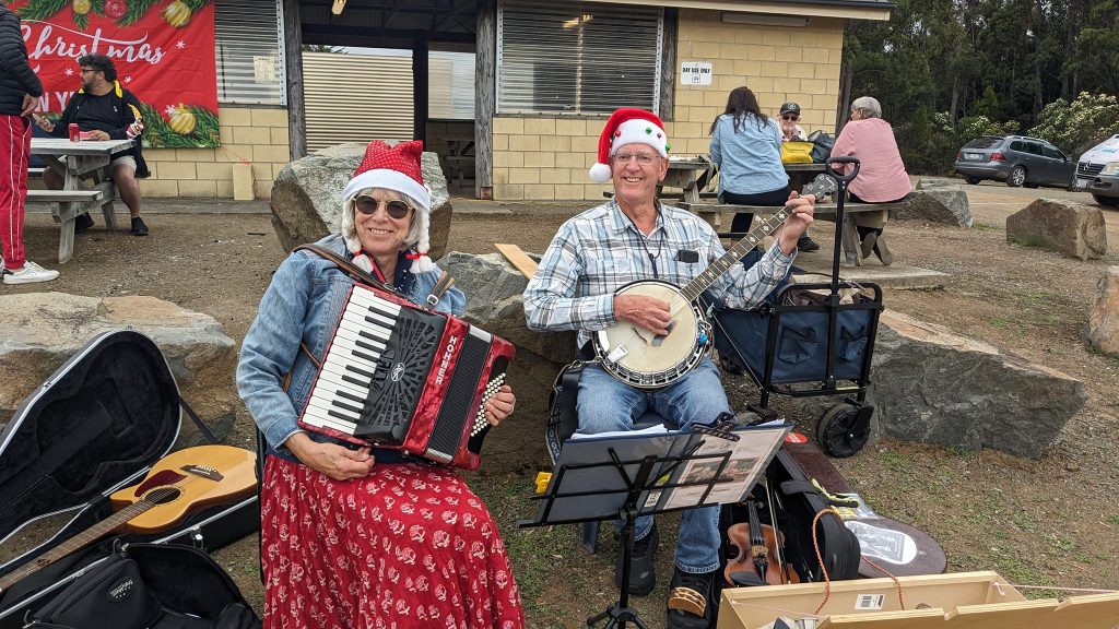 Two people wearing Santa hats play instruments outdoors; the woman plays an accordion and the man plays a banjo. People sit at picnic tables in the background, and musical equipment is scattered around them.