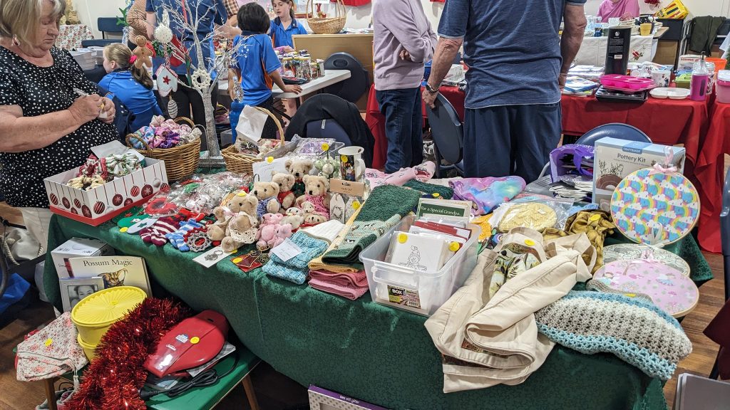 A busy indoor market table displays handmade crafts, stuffed animals, towels, baskets, books, and festive decorations. People browse and chat in the background.