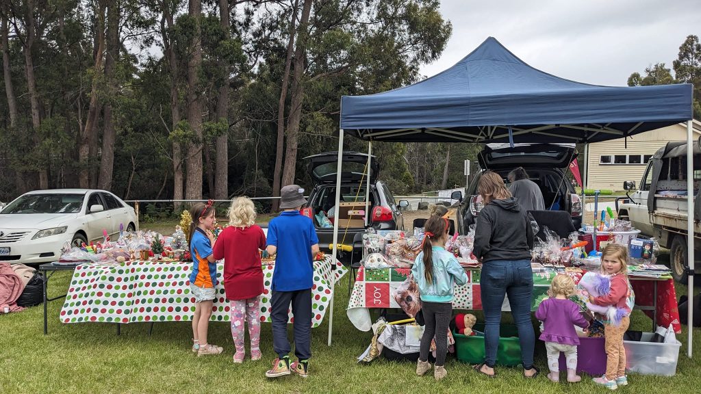 Children and adults gather at a market stall under a blue canopy, with tables covered in treats and gifts. Cars are parked nearby, and the scene is set on grass with trees and a building in the background.
