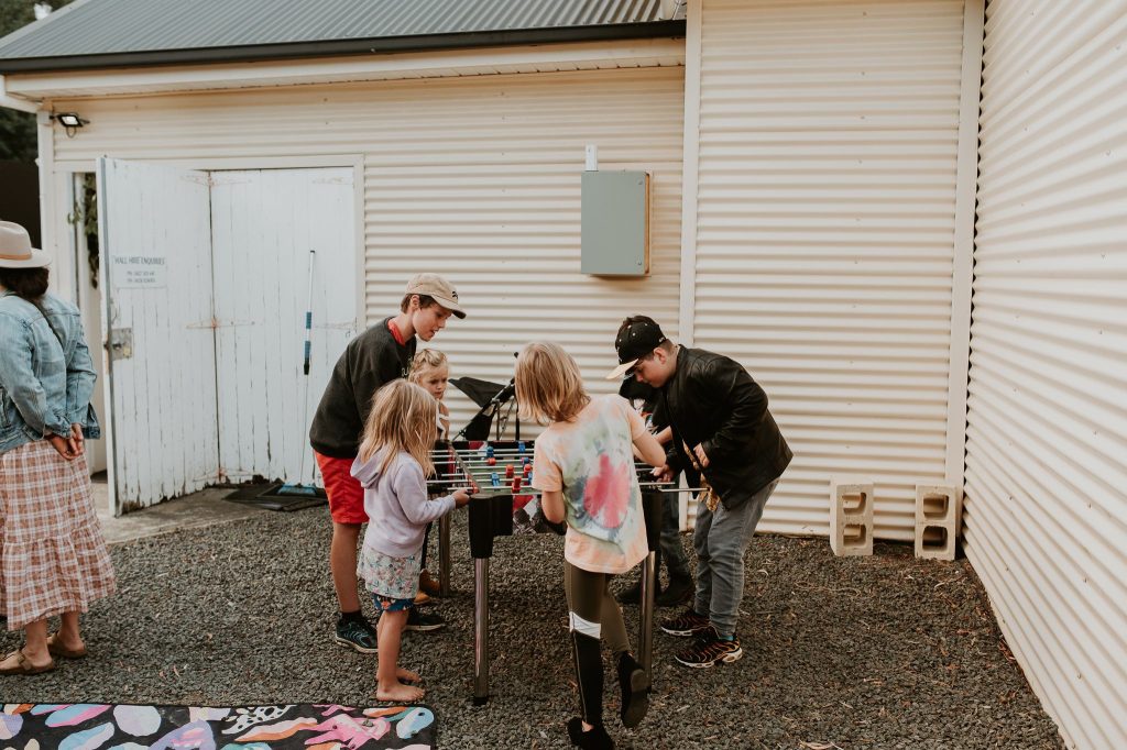 A group of children and one adult stand outside by a foosball table, playing together on a gravel area next to a cream corrugated metal building. One woman stands nearby watching, wearing a hat and plaid skirt.