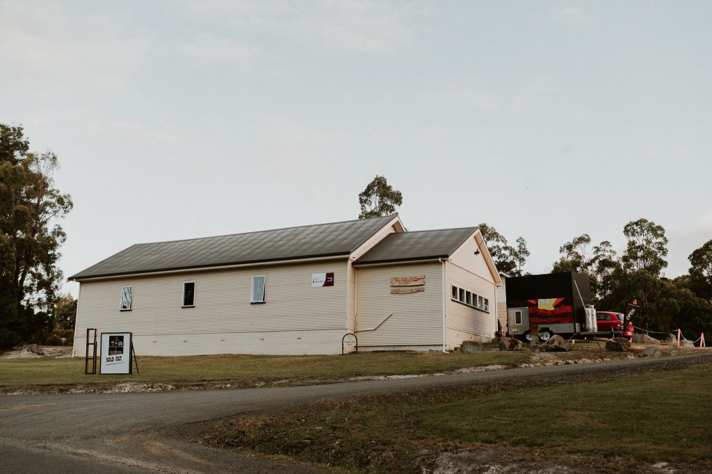 A beige, single-story building with a gray roof sits on a grassy area beside a paved road. There are signs and banners on the building and a small shed with a mural nearby. Trees are in the background.