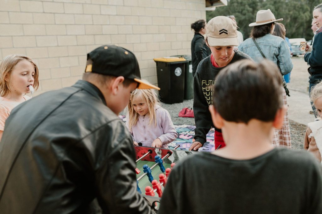 A group of children and adults gather outdoors, with several kids playing foosball on a table. Others watch or interact nearby. There is a yellow-lidded trash bin and a brick wall in the background.