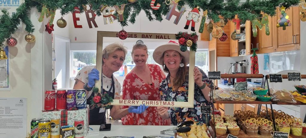 Three smiling women stand behind a counter of festive treats and drinks, holding a Merry Christmas frame. Christmas decorations and a garland hang above them in a cheerful indoor setting.