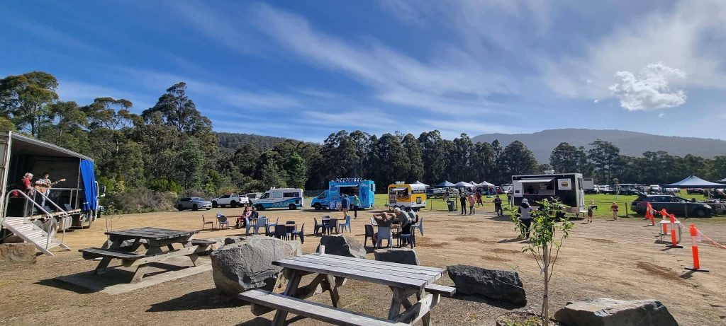 Outdoor food truck event with picnic tables, scattered chairs, people sitting and walking, several food trucks lined up, surrounded by trees and mountains under a blue sky with wispy clouds.