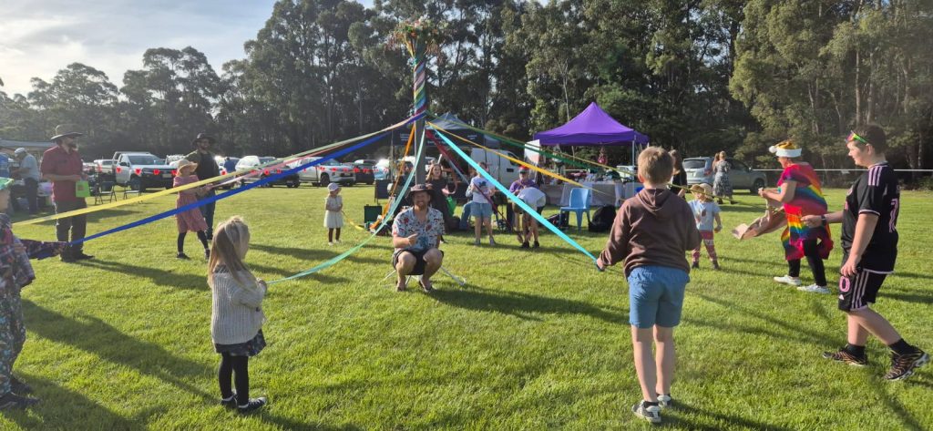 A group of people, including children and adults, dance around a maypole with colorful ribbons in a grassy park, surrounded by trees, parked cars, and a purple tent in the background.