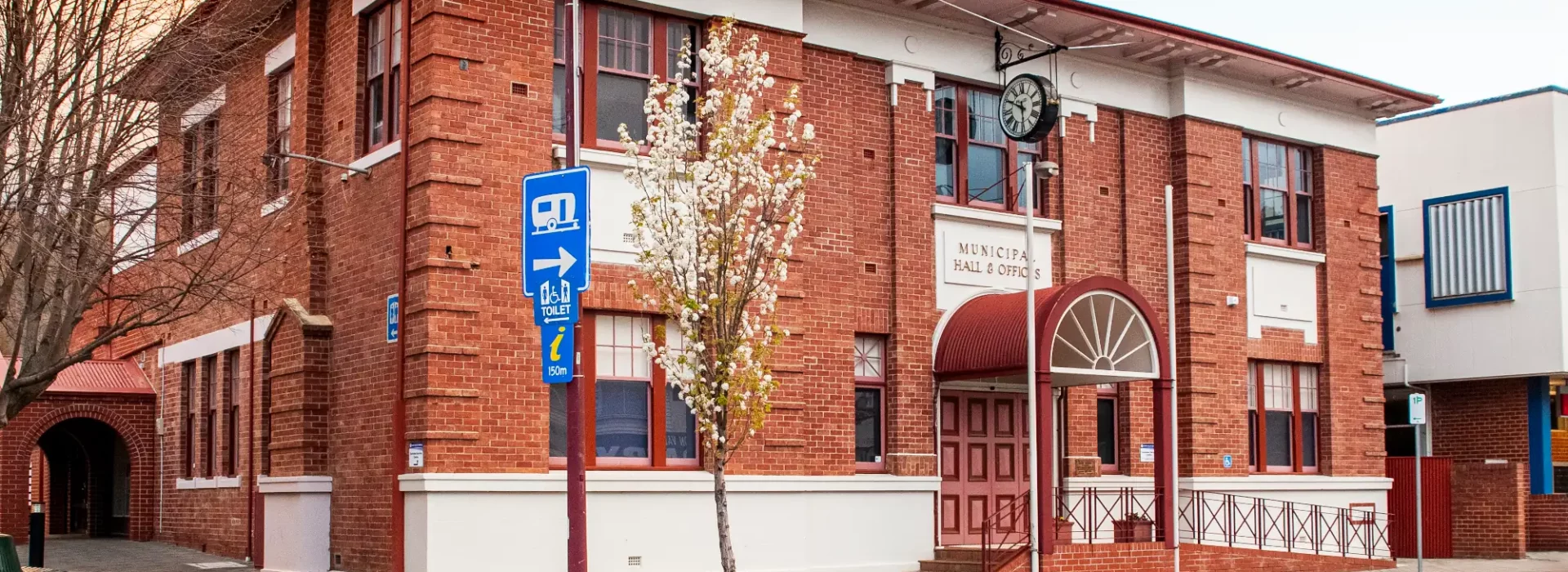 A red brick municipal building with arched windows, a clock above the entrance, and a white and maroon color scheme sits on a quiet street. A single tree with yellow leaves grows nearby under a partly cloudy sky.