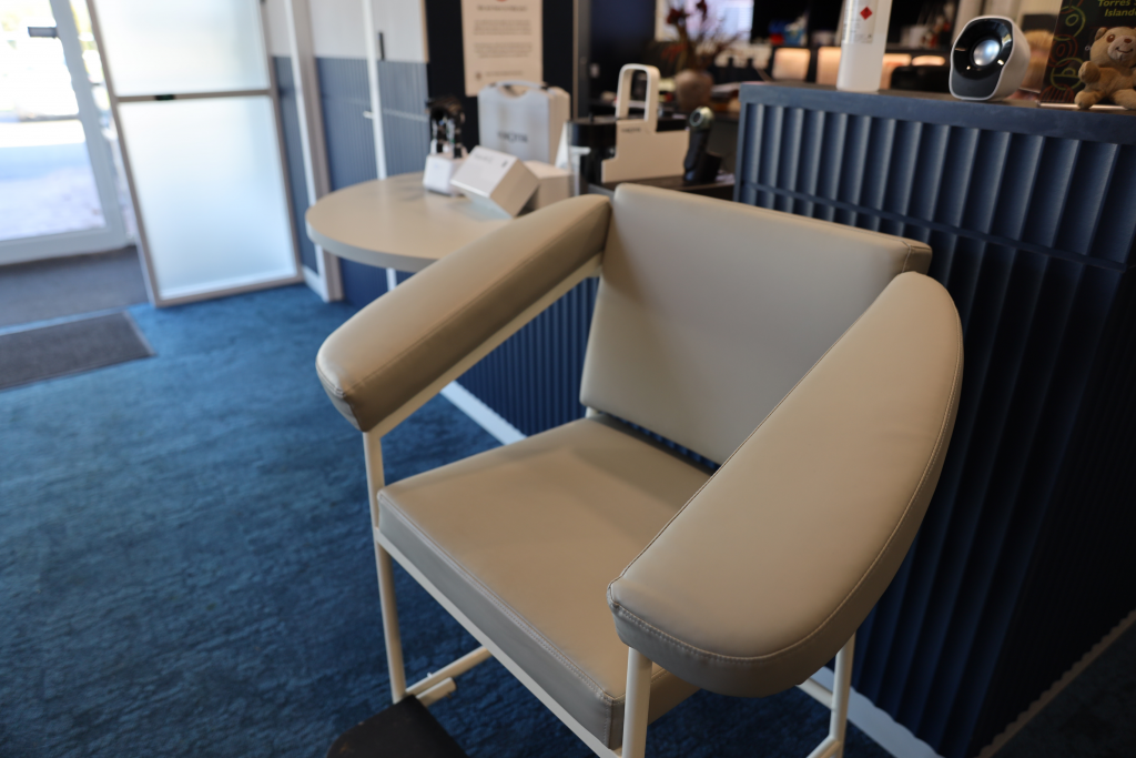 A padded light gray chair with wide armrests is positioned on a blue carpet in a modern office or clinic, next to a round table and dark blue paneled wall with electronic devices and brochures displayed.