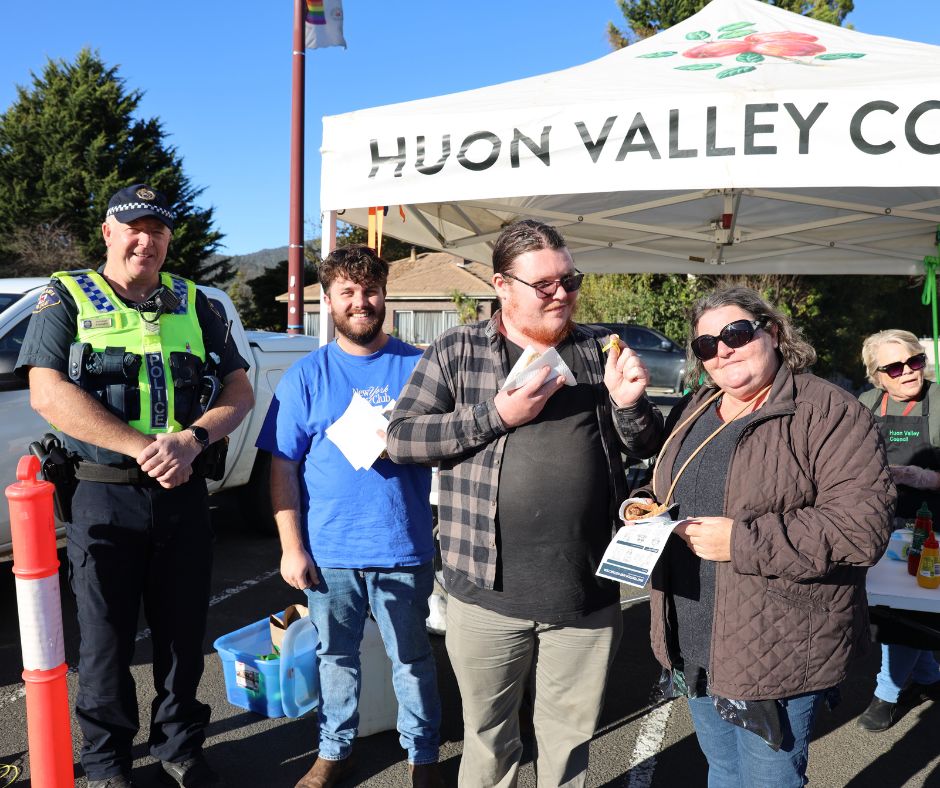 Four people stand in front of a Huon Valley Council tent outdoors, smiling at the camera. One is a police officer in uniform, while the others are casually dressed, holding leaflets and snacks. It is a sunny day.