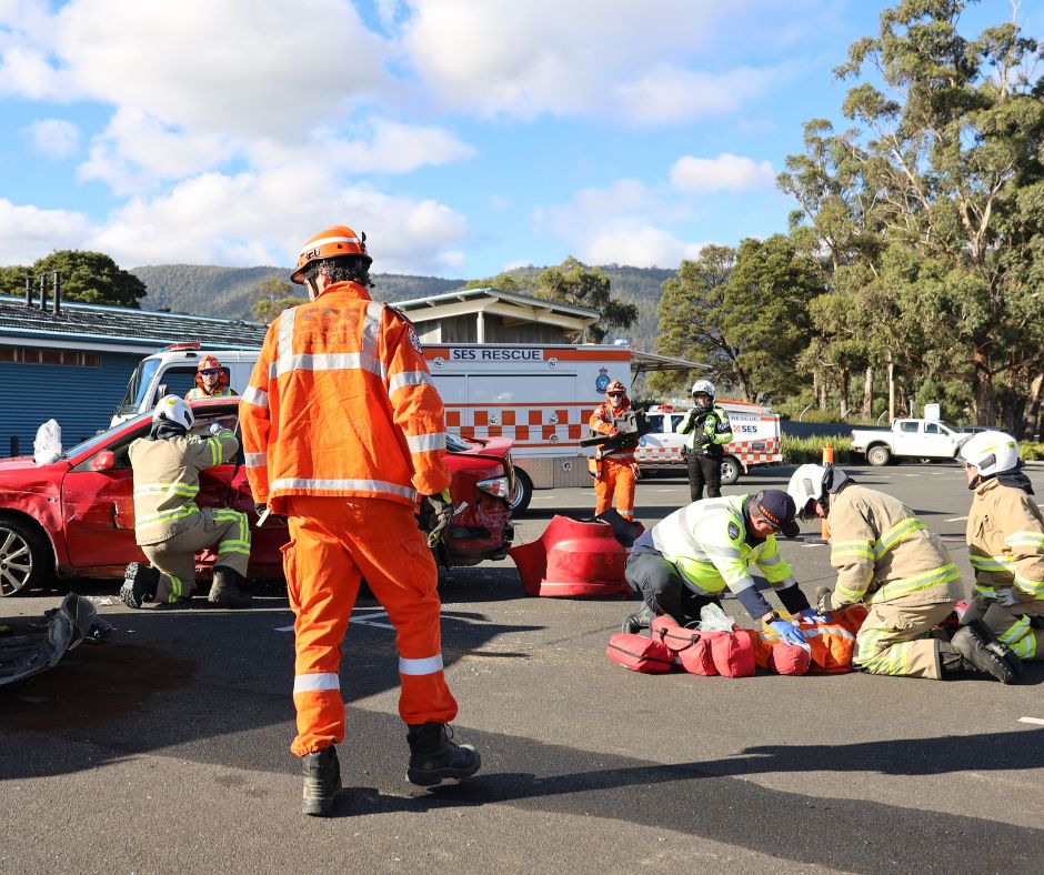 Emergency responders in orange and yellow uniforms assist at the scene of a car accident, with a red vehicle and rescue equipment visible. An ambulance and rescue vehicle are parked nearby under a partly cloudy sky.