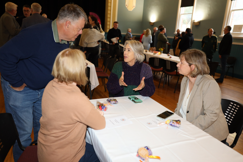 Four adults converse around a table covered with a white cloth at a community event in a hall. Other people mingle in the background. Two women are seated, one man and one woman are standing, all engaged in conversation.