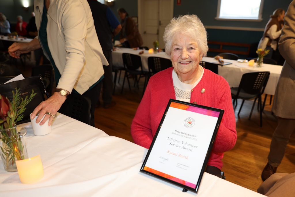 An elderly woman with white hair and a pink sweater smiles while holding a framed Lifetime Volunteer Service Award at a table in a brightly lit hall. Other people are gathered and seated in the background.