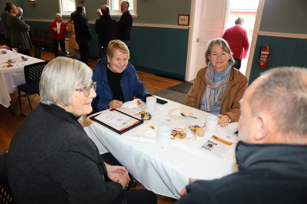 Four older adults sit around a table with coffee cups and plates, talking and smiling in a brightly lit room set up for a gathering. Other people stand and chat in the background near an open door.