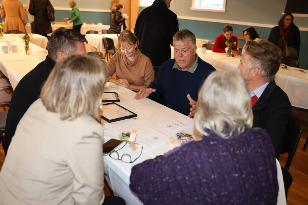 A group of six adults sit around a table covered with a white tablecloth, engaged in conversation at a social gathering in a community hall. Other people and tables are visible in the background.
