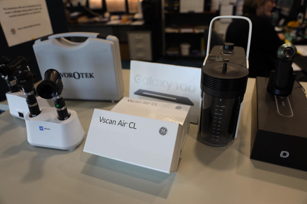 A display of medical and electronic devices on a table, including a Vscan Air CL box, various diagnostic instruments, a container with measurement markings, and a box labeled Galaxy Tab in the background.