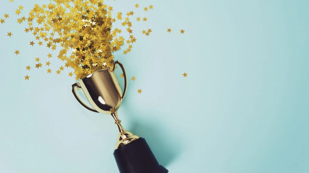 A gold trophy lying on its side with small gold star confetti spilling out of it against a light blue background.