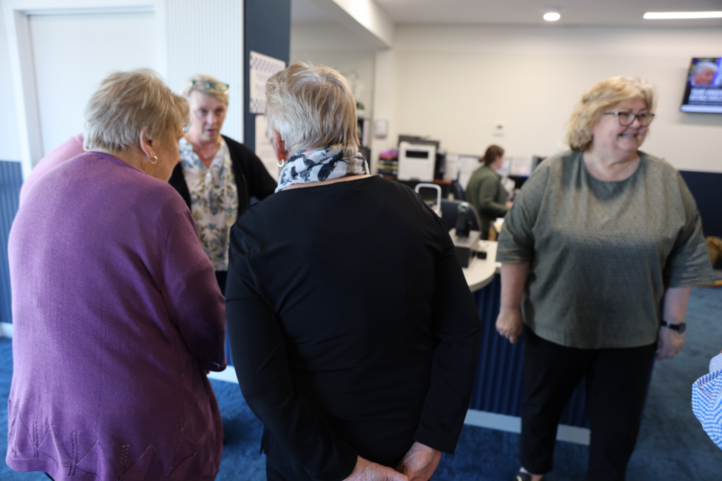 Four women are standing and conversing in an office with blue carpet. Three are in the foreground, seen from behind or side, while one woman stands near a desk and smiles. Office equipment and a TV screen are visible in the background.
