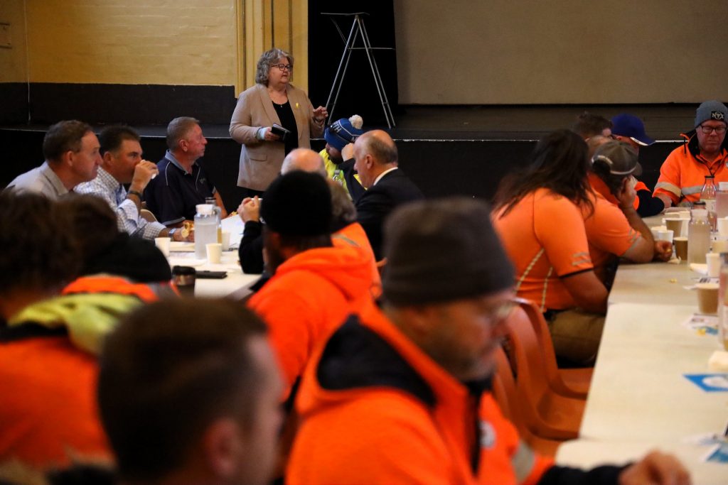 A woman stands speaking to a group of people, many in orange workwear, seated at long tables in a hall. Some have food and drinks in front of them. It appears to be a community or workplace meeting.