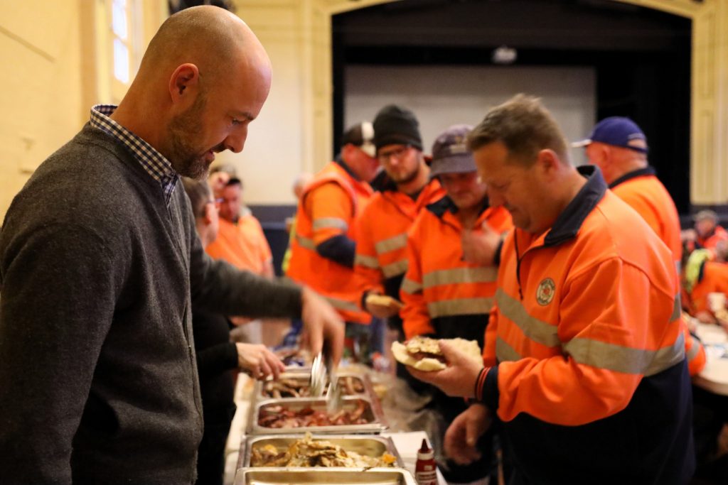 A man serves food from a buffet line to workers wearing orange high-visibility uniforms inside a large indoor hall. The workers hold plates and appear to be waiting their turn.