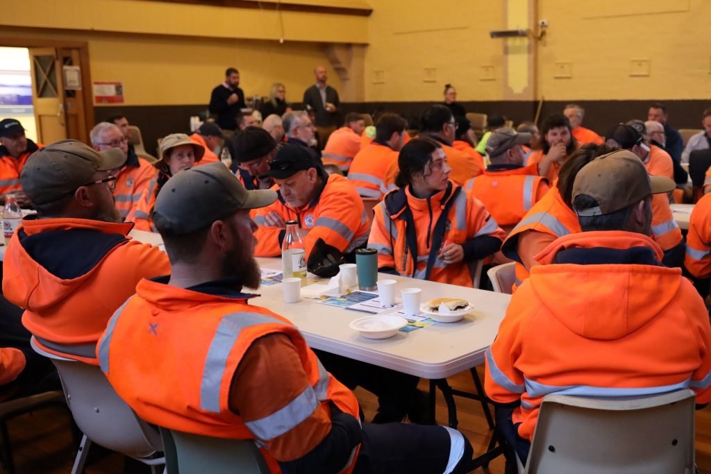 A large group of people in high-visibility orange jackets sit around tables in a community hall, eating and talking. Some stand in the background, and papers and mugs are visible on the tables.