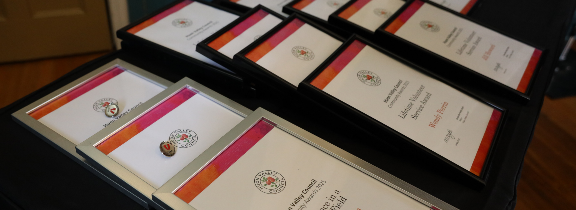 A table displays multiple framed certificates and enamel pins, arranged in neat rows on a black tablecloth, ready for an award ceremony. A blue wall and a door are visible in the background.
