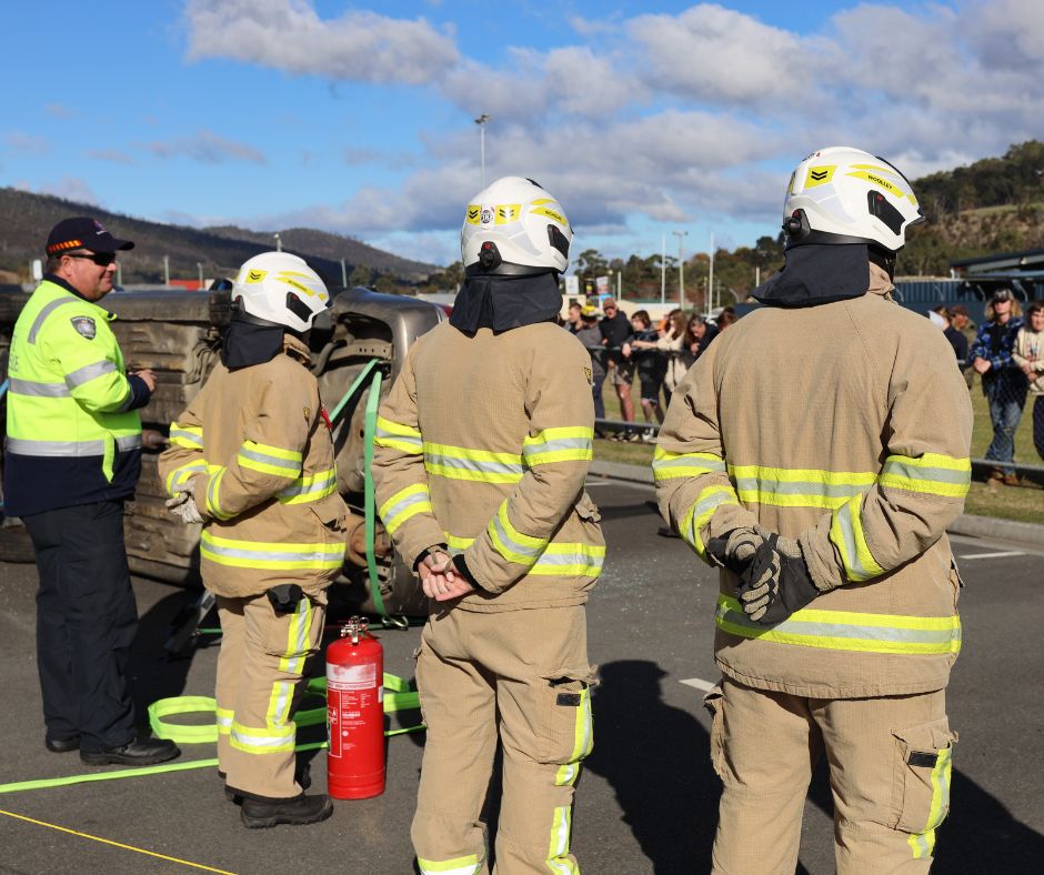 Three firefighters in tan gear and helmets stand near a flipped car, with a fire extinguisher on the ground. A uniformed official is nearby and a crowd watches behind a barrier under a partly cloudy sky.